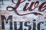A rustic wooden sign with peeling paint features large, stylized lettering in black and red that reads 'Live Music'. The background consists of weathered planks, giving it a vintage and worn appearance.
