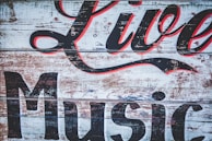A rustic wooden sign with peeling paint features large, stylized lettering in black and red that reads 'Live Music'. The background consists of weathered planks, giving it a vintage and worn appearance.