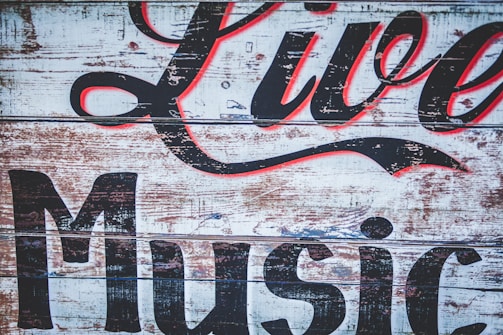 A rustic wooden sign with peeling paint features large, stylized lettering in black and red that reads 'Live Music'. The background consists of weathered planks, giving it a vintage and worn appearance.