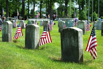 Numerous gravestones are arranged in rows within a cemetery. Each headstone is adorned with a small American flag, creating a memorial atmosphere. The surrounding area is lush with green grass and tall trees, and other flags can be seen in the distance.