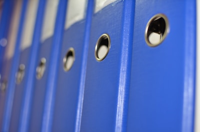 A row of blue office binders is aligned on a shelf, each featuring a circular metallic ring for easy handling. The focus is on the binders, with a perspective that captures their uniformity and professional appearance.