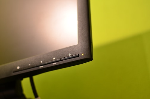 A close-up view of a computer monitor's lower edge, highlighting several control buttons, including a power button with an indicator light. The background is a solid, bright green, which contrasts with the dark frame of the monitor.