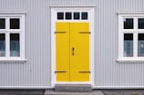 A building featuring a bright yellow door with black hinges and a handle, set within a gray corrugated metal facade. White-trimmed windows flank the door on both sides, with decorative black ironwork details on the glass.