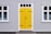 A building featuring a bright yellow door with black hinges and a handle, set within a gray corrugated metal facade. White-trimmed windows flank the door on both sides, with decorative black ironwork details on the glass.