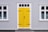A building featuring a bright yellow door with black hinges and a handle, set within a gray corrugated metal facade. White-trimmed windows flank the door on both sides, with decorative black ironwork details on the glass.