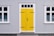 A building featuring a bright yellow door with black hinges and a handle, set within a gray corrugated metal facade. White-trimmed windows flank the door on both sides, with decorative black ironwork details on the glass.