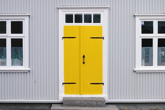 A building featuring a bright yellow door with black hinges and a handle, set within a gray corrugated metal facade. White-trimmed windows flank the door on both sides, with decorative black ironwork details on the glass.