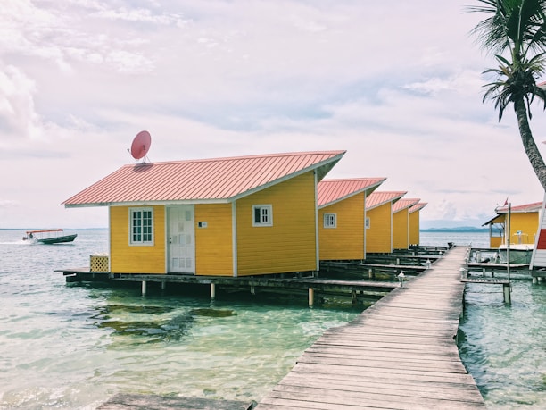 A series of vibrant yellow bungalows with red roofs sit on stilts above clear turquoise waters, connected by a wooden walkway. A palm tree and a boat in the distance add to the tropical setting.