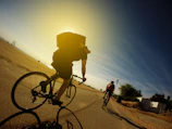 Cyclist riding along a sunlit trail with rolling green hills and a glimpse of the sea in the background.