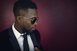 A person wearing a suit and tie is looking to their left. They have close-cropped hair and are wearing stylish sunglasses. The background is a deep, solid color.