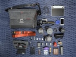A neatly arranged flat lay of photography equipment and accessories on a striped surface. Items include a grey fabric camera bag with a shoulder strap, an array of camera lenses, a digital camera, and several small gadgets like a phone, a portable battery, USB cables, and various camera accessories. A tablet and earphones are also present, with a red camera strap and other small items like SD cards and a lens cleaning tool.