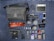 A neatly arranged flat lay of photography equipment and accessories on a striped surface. Items include a grey fabric camera bag with a shoulder strap, an array of camera lenses, a digital camera, and several small gadgets like a phone, a portable battery, USB cables, and various camera accessories. A tablet and earphones are also present, with a red camera strap and other small items like SD cards and a lens cleaning tool.