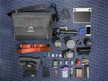 A neatly arranged flat lay of photography equipment and accessories on a striped surface. Items include a grey fabric camera bag with a shoulder strap, an array of camera lenses, a digital camera, and several small gadgets like a phone, a portable battery, USB cables, and various camera accessories. A tablet and earphones are also present, with a red camera strap and other small items like SD cards and a lens cleaning tool.
