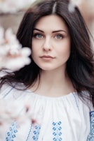 A woman with long dark hair and striking eyes is wearing a white blouse with blue embroidery. The background features soft-focus flowers, creating a serene and natural setting.
