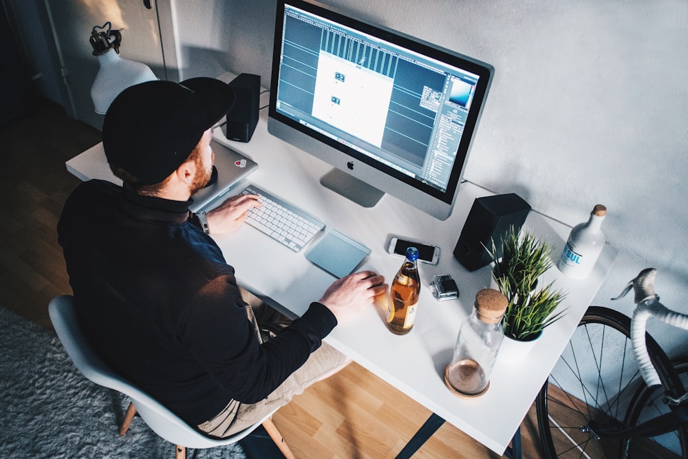 A man wearing a black longsleeve shirt and black hat, using a Mac desktop computer.