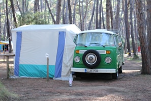 A vintage green camper van is parked next to a white and blue tent in a forested campsite. The area is surrounded by tall pine trees, creating a serene and natural setting. The van has a spare tire mounted on the front and appears to be set up for camping, with gear likely inside.