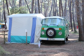 A vintage green camper van is parked next to a white and blue tent in a forested campsite. The area is surrounded by tall pine trees, creating a serene and natural setting. The van has a spare tire mounted on the front and appears to be set up for camping, with gear likely inside.