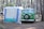 A vintage green camper van is parked next to a white and blue tent in a forested campsite. The area is surrounded by tall pine trees, creating a serene and natural setting. The van has a spare tire mounted on the front and appears to be set up for camping, with gear likely inside.