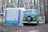 A vintage green camper van is parked next to a white and blue tent in a forested campsite. The area is surrounded by tall pine trees, creating a serene and natural setting. The van has a spare tire mounted on the front and appears to be set up for camping, with gear likely inside.