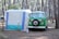 A vintage green camper van is parked next to a white and blue tent in a forested campsite. The area is surrounded by tall pine trees, creating a serene and natural setting. The van has a spare tire mounted on the front and appears to be set up for camping, with gear likely inside.