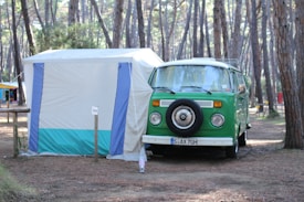 A vintage green camper van is parked next to a white and blue tent in a forested campsite. The area is surrounded by tall pine trees, creating a serene and natural setting. The van has a spare tire mounted on the front and appears to be set up for camping, with gear likely inside.