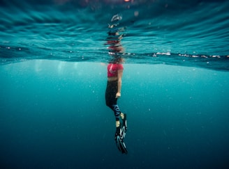 A person is partially submerged in water, with the body below the surface and the head above. Wearing a pink top and black leggings, they have fins on their feet. The water is clear, with a bluish tint, and there is a reflective rippling effect on the surface.