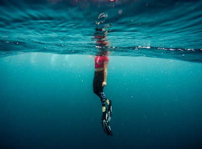 A person is partially submerged in water, with the body below the surface and the head above. Wearing a pink top and black leggings, they have fins on their feet. The water is clear, with a bluish tint, and there is a reflective rippling effect on the surface.