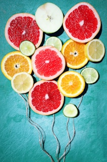Slices of various citrus fruits including grapefruit, lemon, lime, and orange are arranged creatively on a bright teal background. Each slice is vivid and fresh, showing a range of colors and textures. Strings are attached to the slices, making them resemble colorful balloons.