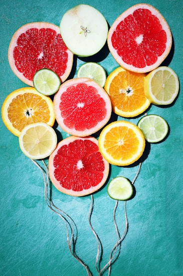 Slices of various citrus fruits including grapefruit, lemon, lime, and orange are arranged creatively on a bright teal background. Each slice is vivid and fresh, showing a range of colors and textures. Strings are attached to the slices, making them resemble colorful balloons.