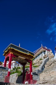 An ornate, traditional archway painted in red and gold stands prominently against a bright blue sky. Behind the archway, there is a stone pathway that leads to a monastery situated on a rocky hill. The monastery's buildings are decorated with vibrant prayer flags.