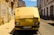 A yellow van with cardboard covering the windshield, parked on a narrow cobblestone street lined with old, weathered buildings. Graffiti is visible on the left wall, and the van bears a license plate marking it as registered in Lisbon.