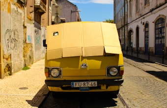 A yellow van with cardboard covering the windshield, parked on a narrow cobblestone street lined with old, weathered buildings. Graffiti is visible on the left wall, and the van bears a license plate marking it as registered in Lisbon.