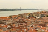 A picturesque view of a coastal cityscape featuring a variety of buildings with red-tiled roofs. In the background, a wide river is visible, crossed by a large suspension bridge. A prominent monument stands on a distant hillside across the river.