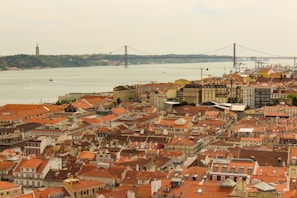 A picturesque view of a coastal cityscape featuring a variety of buildings with red-tiled roofs. In the background, a wide river is visible, crossed by a large suspension bridge. A prominent monument stands on a distant hillside across the river.