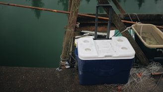 A large rectangular cooler with a blue bottom and white top sits on a wooden dock by the water. It has a handle for carrying and is surrounded by wooden beams, a net, and another storage container. The scene suggests a fishing or boating area.