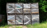 A stack of wooden crates, each labeled with 'Lakeville Orchards', is placed against a backdrop of dense green foliage. The crates, with visible wear and some faded paint, are arranged in a neat grid pattern. Sunlight filters through the trees, casting dappled shadows on the crates and the ground, which is covered with grass.