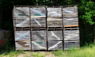 A stack of wooden crates, each labeled with 'Lakeville Orchards', is placed against a backdrop of dense green foliage. The crates, with visible wear and some faded paint, are arranged in a neat grid pattern. Sunlight filters through the trees, casting dappled shadows on the crates and the ground, which is covered with grass.