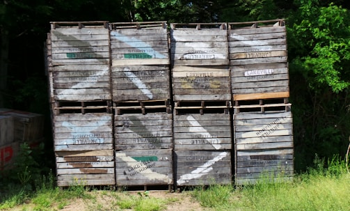 A stack of wooden crates, each labeled with 'Lakeville Orchards', is placed against a backdrop of dense green foliage. The crates, with visible wear and some faded paint, are arranged in a neat grid pattern. Sunlight filters through the trees, casting dappled shadows on the crates and the ground, which is covered with grass.