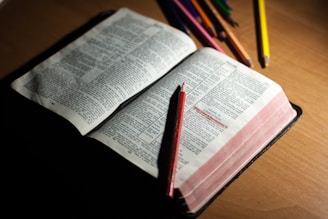 An open book with visible text, possibly a Bible or similar text, is laying on a wooden surface. Several colored pencils are scattered around, with one red pencil resting on the open pages.
