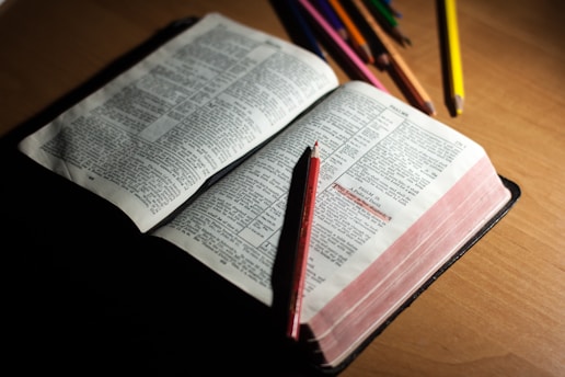 An open book with visible text, possibly a Bible or similar text, is laying on a wooden surface. Several colored pencils are scattered around, with one red pencil resting on the open pages.