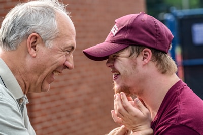 An elderly man and a younger man wearing a maroon cap and shirt share a moment of laughter outdoors against a brick wall backdrop. The close-up image captures expressions of joy and connection.
