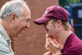 An elderly man and a younger man wearing a maroon cap and shirt share a moment of laughter outdoors against a brick wall backdrop. The close-up image captures expressions of joy and connection.