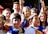 A group of young people are gathered together, several with painted faces in blue, yellow, and red colors. They are smiling and appear to be enjoying themselves. Flags or similar paraphernalia are draped on their shoulders or held in their hands. The scene is bright and lively, set outdoors.