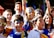 A group of young people are gathered together, several with painted faces in blue, yellow, and red colors. They are smiling and appear to be enjoying themselves. Flags or similar paraphernalia are draped on their shoulders or held in their hands. The scene is bright and lively, set outdoors.