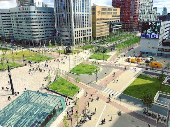 An urban landscape featuring a busy intersection surrounded by modern high-rise buildings and a public square. There is a large glass structure in the foreground, numerous pedestrians walking, and some cyclists on the pathways. Green spaces with neatly arranged trees add a touch of nature to the cityscape.