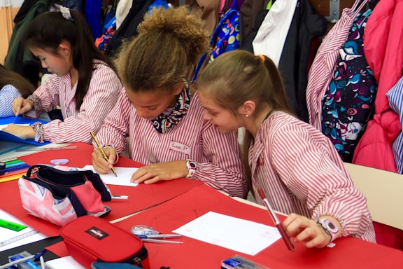Children wearing striped uniforms sit at a table covered with red and white paper, engaging in an art or writing activity. Colored pencils, pens, and pouches are scattered across the table, while coats and colorful backpacks hang in the background.