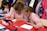 Children wearing striped uniforms sit at a table covered with red and white paper, engaging in an art or writing activity. Colored pencils, pens, and pouches are scattered across the table, while coats and colorful backpacks hang in the background.