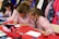 Children wearing striped uniforms sit at a table covered with red and white paper, engaging in an art or writing activity. Colored pencils, pens, and pouches are scattered across the table, while coats and colorful backpacks hang in the background.