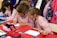 Children wearing striped uniforms sit at a table covered with red and white paper, engaging in an art or writing activity. Colored pencils, pens, and pouches are scattered across the table, while coats and colorful backpacks hang in the background.