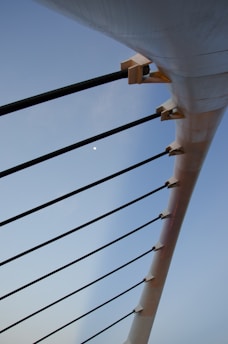 A close-up view of the structural elements of a bridge or similar architecture, featuring long black support cables attached to a large white beam, with a clear blue sky in the background.